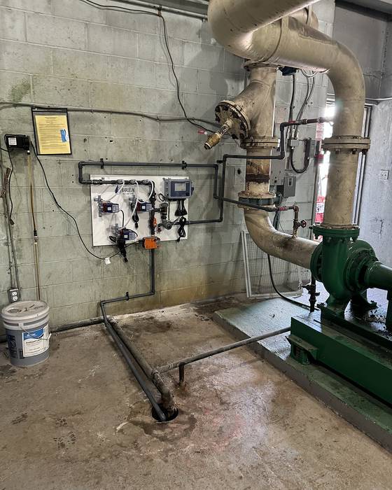 Wall-mounted chemical feed panel above a floor drain, with a green base-mounted pump and large piping