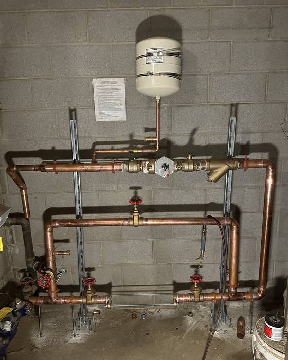 Copper piping assembly with a small expansion tank and red isolation valves on a block wall