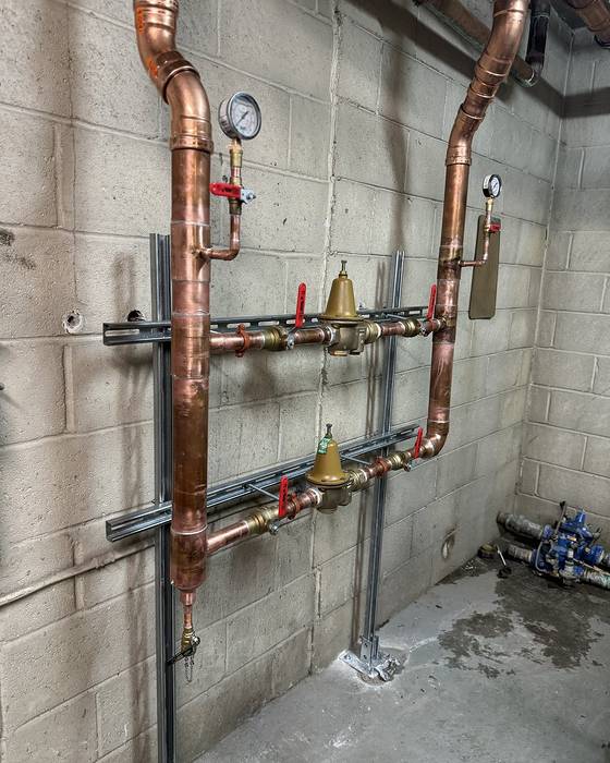 Two brass pressure-reducing valves with gauges on copper piping mounted to strut on a block wall