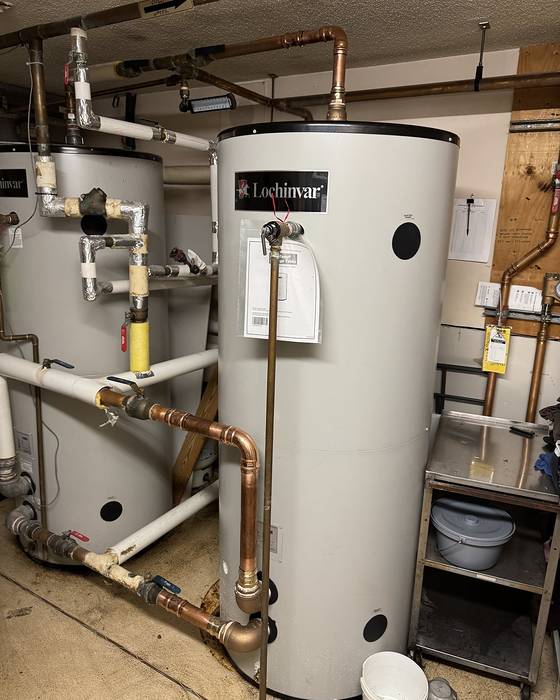 Two tall storage water heaters with copper and white piping in a utility room