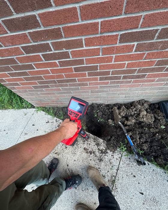 Technician using a red utility locator camera beside a brick wall while digging by hand