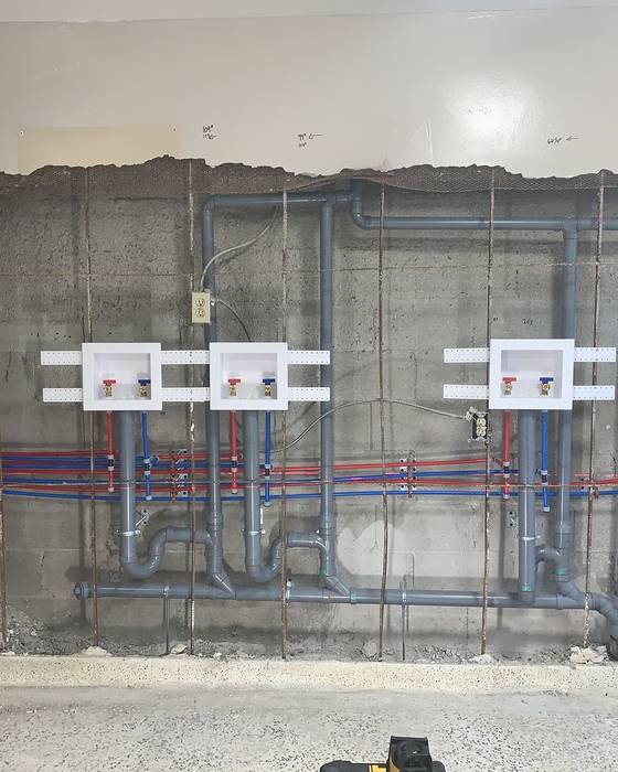 Rough-in of four washing machine outlet boxes with red and blue PEX supply lines and grey drain piping on a concrete wall